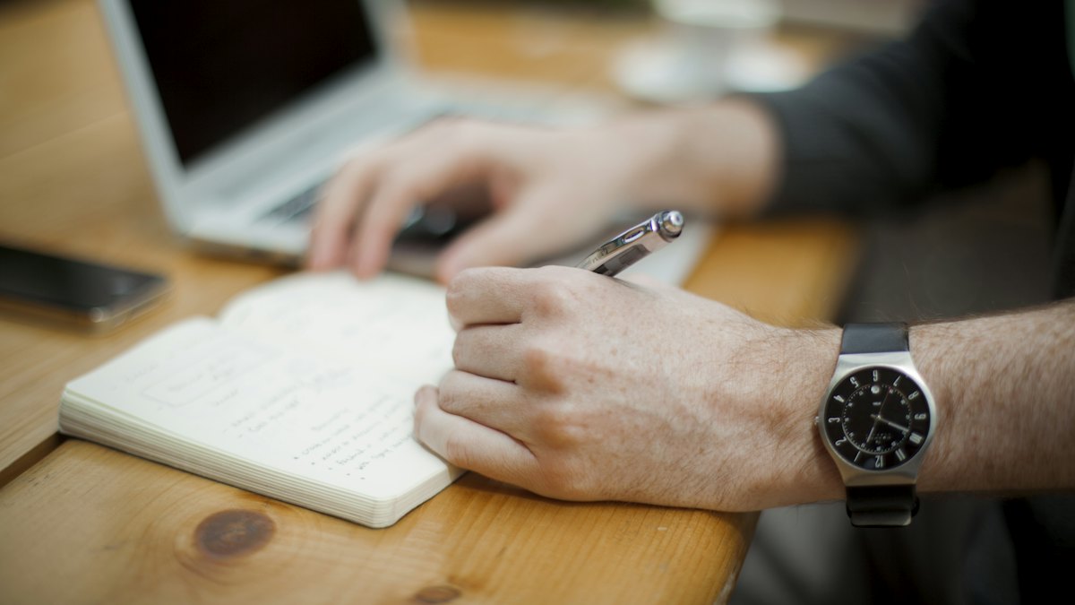 Main qui écrit dans un carnet ouvert posé sur un bureau en bois, devant un ordinateur portable, pour s'exercer à reformuler