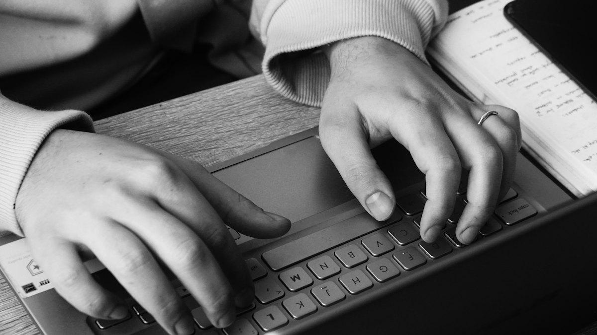 Deux mains qui tapent sur le clavier d'un ordinateur portable, un carnet de notes posé à côté